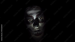 Portrait of creepy woman with a make-up for Halloween in santa muerte style illuminated by red lights. A lady with painted face in close-up looks steadily and bends slowly her head on black background
