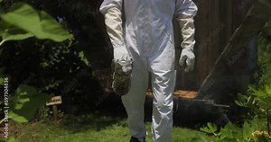 Beekeeper in protective suit holding bee smoker and walking in slow motion at beehive at the apiary.