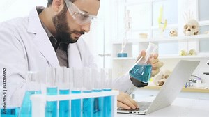 Caucasian scientist holding test tube for analyzing and using laptop for note experiment data.