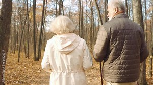 Sporty active retired beautiful man hiking walking slowly in autumn park with grey-haired senior pretty woman. Good-looking elderly female doing sports activities in city with older bearded husband.