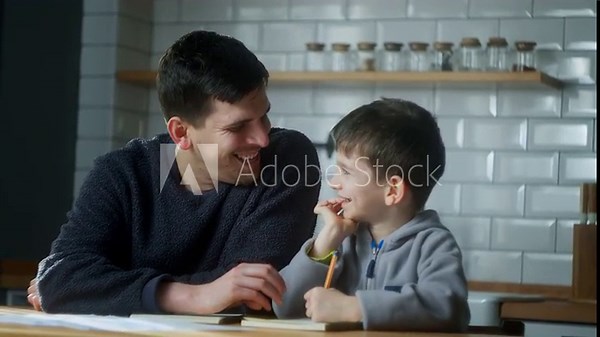 Young father help little schoolboy pupil with homework at home. Dad helping to son with writing while sitting in the kitchen. Adult parent or tutor with kid boy learning. Private education