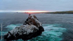 Monolithic bird rookery rock, covered in white guano, drone pulls back, sunset in Pacific Ocean, Central California Coast with Piedras Blancas Lighthouse in distance, crashing waves, 4k Pro Res 422HQ