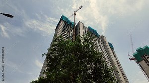 Low-angle view of a high-rise residential building under construction against a vivid blue sky. Towering structure with exposed framework and scaffolding. Cranes visible at the top.
