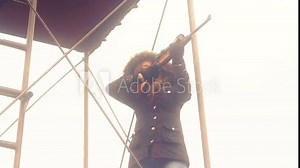 A sneaky view behind bushes of a woman taking aim with a shotgun on top of a metal tower fort, 30 fps.