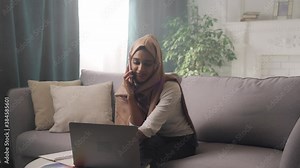 Positive successful Indian woman wearing hijab scarf working from home, multitasking, using laptop, typing, having a call, speaking to clients on phone, enjoying remote job.