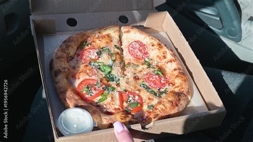 A person is holding a pizza box open to reveal a freshly made pizza inside, featuring sliced tomatoes and toppings. The setting is the interior of a vehicle, and sunlight is coming through the window