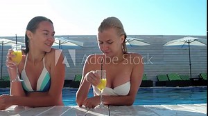 Two happy girlfriends having fun in the pool. close to the side of the pool, takes an alcoholic drink, takes a sip of alcohol through a straw, expensive vacation in the resort pool.