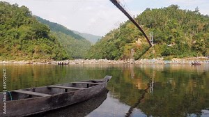 close up shot of a stranded or Docked Wooden fishing boat in lake or pond while a group of paddlers propelling a canoe in background with bridge and lush green trees in mountainous region in daylight.