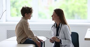 Young woman doctor pediatrician in white uniform ask illness symptoms, writes, filling form talks to little 8s boy patient during visit in clinic or hospital. Medicine, paediatric, health diagnostics
