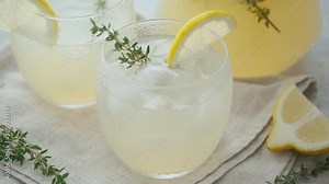 Refreshing lemonade with thyme in a transparent glass and a jug with lemonade on the background. Cold summer drink with lemon and thyme. Summer refreshing cocktail concept. slider shot 4K UHD