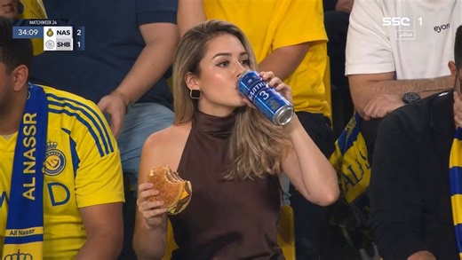 Made with seedance 2.0 + GPT Image 2 Prompt: Ultra-realistic sports broadcast still of a glamorous woman sitting in a packed football stadium crowd during a night match, wearing a dark brown sleeveless high-neck satin top and black square earrings, shoulder-length light brown/blonde hair styled in soft waves. She is casually drinking from a tall blue aluminum can while holding a half-eaten cheeseburger in the other hand. Around her are fans in bright yellow and blue football jerseys and scarves,