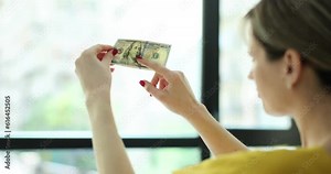Woman checks watermarks on money dollars in front of window. Recognize counterfeit banknotes at home