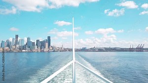 Steady view of Downtown Seattle, CenturyLink field, T-Mobile Park, and Port of Seattle from water off stern of Washington State Ferry. Wake is seen behind steering pole on sunny summer day.