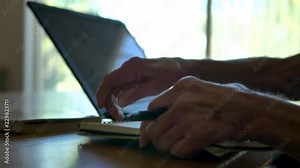 An elderly woman with wrinkled hands writes with a pen in a notebook as she look on her laptop backlit by a bright window.
