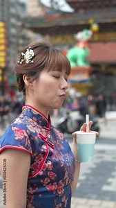 A vertical video of a Taiwanese woman in her 20s wearing a blue traditional Chinese dress in Wanhua District, Taipei City, Taiwan, walking around the town near Longshan Temple while drinking tapioca m
