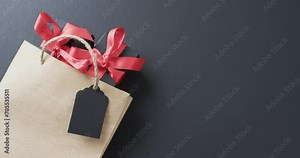 A brown paper gift bag with a vibrant red ribbon and a blank tag lies on a dark surface, with copy s