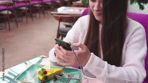 Beautiful young girl with long hair chatting on mobile phone with friend. Woman write message on smartphone in cafe and smiling on background with yellow cake and pink drink