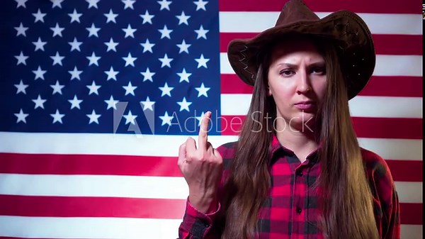 Woman with long hair in a cowboy hat and a checkered black and red shirt on a background of the American flag, shows an indecent sign, a phallic symbol.