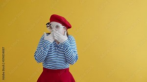 Young mime woman closing her mouth with both hands in gloves showing her extreme surprise. Mime playing funny emotional performance on yellow background