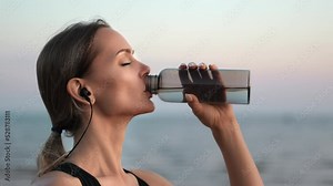 Athletic runner woman in earphones drinking pure water bottle training at sea coast sunset beach