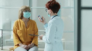 Female doctor in mask scanning forehead of senior woman with infrared thermometer and writing temperature measurements on clipboard during medical checkup in clinic in time of covid-19 pandemic