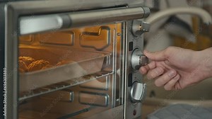 Side stab closeup of hand turning power control and timer control on toaster oven with baking tray of biscuits inside in modern kitchen
