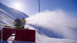 Snow cannon at ski resort in winter, snow machine produced artificial snow in mountains on slope at sunny day. Preparation of ski track in hills for active sport. Turbine spray water in cold weather.