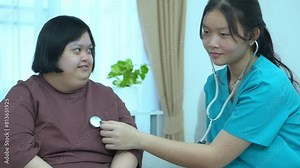 Asian female nurse uses stethoscope to perform physical examination of woman with Down syndrome at home.