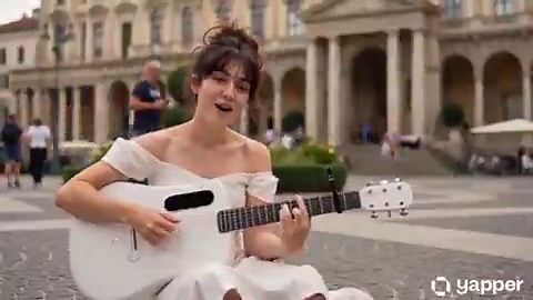 Seedance 2.0 on @yapper_soPrompt:Cinematic, high-quality video of a beautiful young woman with auburn hair in a messy romantic updo, wearing an elegant, flowy, off-the-shoulder white dress. She is sitting gracefully on the cobblestone ground of a sunlit, grand European piazza, gently strumming a white acoustic guitar and singing a song. A fluffy white Ragdoll cat with a bushy tail is walking around her and affectionately nuzzling her leg. The background features classical baroque architecture, a