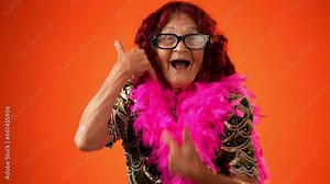 Funny portrait of smiling happy elderly senior old woman with wrinkled skin smiling pointing to camera and giving call me gesture with hands isolated on solid orange background.