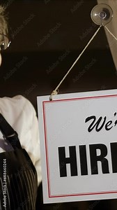 Vertical video of female shop owner putting Help Wanted sign on glass door. Woman staff chef of restaurant advertising for hiring staff.