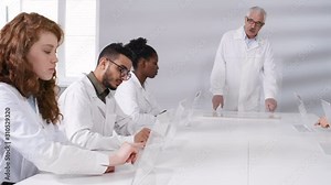 Medium panning shot of senior teacher in lab coat standing in front of multiethnic students, demonstrating basic touchscreen hand gestures, all repeating on futuristic devices. Suitable for AR, VR.