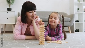 Smiling family woman with wooden brick playing strategy game with tween girl on warm floor of living room. Cheerful mother and daughter building decision-making skills while creating toy skyscraper.