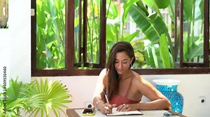 Spanish hipster girl writing informative notes in education textbook using cellphone application at table, young female blogger creating publication text ideas checking smartphone website