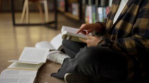 Close-up of a girl student in a plaid shirt reading a book while sitting on the floor among books in the library | Premium Stock Video Footage