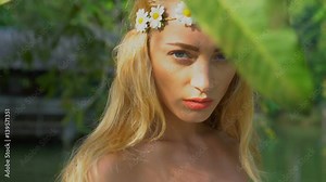 Closeup face of stunning blond woman in white dress and wreath of white flowers looking at the camera and posing with mango fruit tree in the garden over lake background - video in slow motion