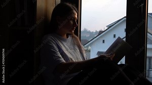 Girl with novel book in hands reads while sitting by opened window on sill. Female reader in cozy home at reading hobby. Rest with literature stress relief. Woman sits on windowsill with drama story. Stock Video