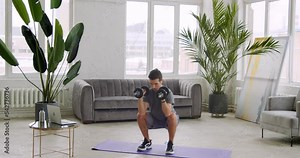 Athletic young man in sportswear doing squats exercise with dumbbells while watching fit training by laptop