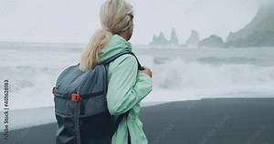 Woman with a green raincoat walking on a black sand beach enjoying view to Reynisfjara sea stack cliff formations close to Vik, Iceland.