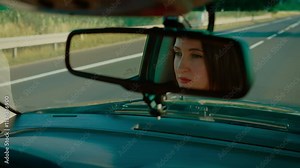 Reflection woman in rearview mirror focusing while driving along highway. Female watching road with concentration framed by mirror. Lady navigating car attentively with highway scenery in background