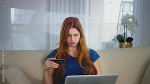 Young Woman Shopping Online with Credit Card. Girl with red hair sits on a couch, holding a credit card and focused on her laptop screen, representing online shopping and digital transactions.