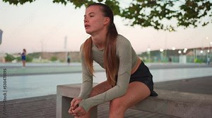 Tired happy woman runner taking a rest after run sitting bench near running track breathe hard. Summer evening time. Young athletic female jogger resting after training. Work hark