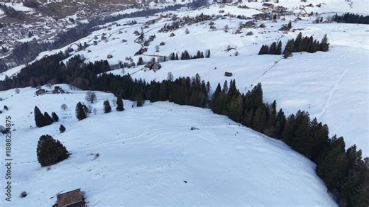 Drone flight forward over a ski slope with skiers descending, alpine cabins and evergreen trees along the piste, small mountain village visible in the distance, winter resort in the Swiss Alps.