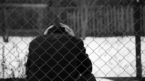 Young unidentifiable teenage boy holding hes head at the correctional institute in black and white, conceptual footage of juvenile delinquency, focus on the wired fence in black and white.