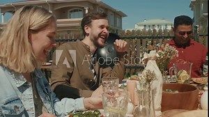 Young Caucasian man with beard on face sitting at table telling funny story to his friends during outdoor party on summer day
