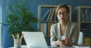 Beautiful Caucasian young lady in glasses sitting at the laptop computer during workday in office and then getting a phone call on her smartphone.
