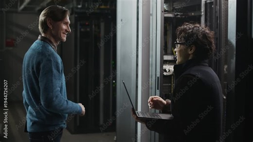 Side view medium shot of two network engineers shaking hands in modern data center corridor after finishing server installation and infrastructure maintenance, one of men continuing work on laptop