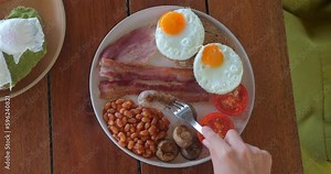Eating sausage using fork. Woman stabs a sausage with a fork while eating. Close up traditional full english breakfast in plate. National British cuisine, fried eggs with bacon, toasts on plate.