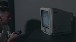 Broken Television or TV Propaganda. Mind Control - Zombified Man in White T-shirt Switches Channels on the Old TV in Dark Room. TV Signal Search. Distortion on the Screen. Medium Close-Up, Film Grain