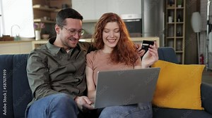 young happy loving couple in home indoors on sofa using laptop computer holding credit card.